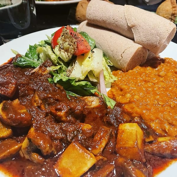 Ethiopian dish with dark stew, orange lentils, fresh salad, and rolled injera bread on a white plate.